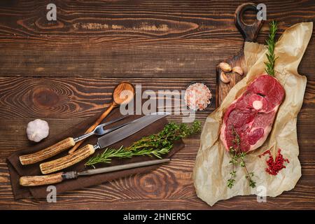 Rohes, frisches Rindfleisch mit mittelgeschnittenem Bein, Gabel, Messer und Spitzer auf Serviette auf braunem Holzhintergrund Stockfoto