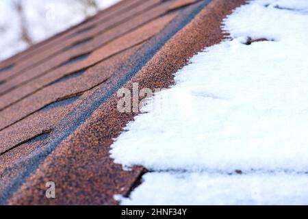 Rote Gürtelrose. Das Dach eines Hauses aus Schindeln ist im Winter mit Schnee bedeckt, Nahaufnahme. Frostbeständigkeit und Elastizität von bituminösen Fliesen in Stockfoto