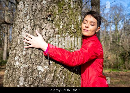 Die junge Frau umarmt eine große Eiche. Die Frau schließt sich und nimmt die Energie der Natur wahr. Konzept von Wohlbefinden und Liebe zur Natur. Stockfoto