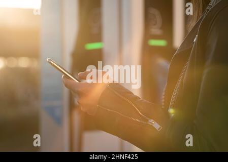 Nahaufnahme der Hand einer Frau, die ein Smartphone neben einer elektrischen Ladestation hält Stockfoto