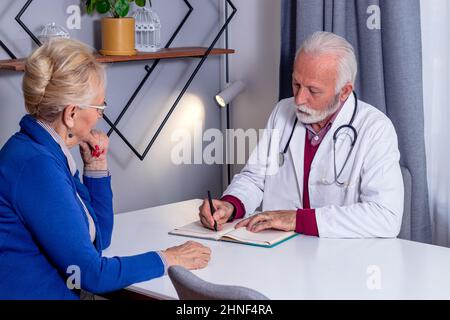 Erfahrener Arzt, der Rezepte für eine ältere Patientin schreibt. Frau mittleren Alters, die ihren Arzt aufgesucht und sich über ihre Krankenversicherung beraten hat. Stockfoto