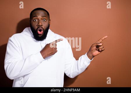 Foto von lustig schockiert Kerl gekleidet weißen Sweatshirt zeigt Finger leeren Raum isoliert braune Farbe Hintergrund Stockfoto
