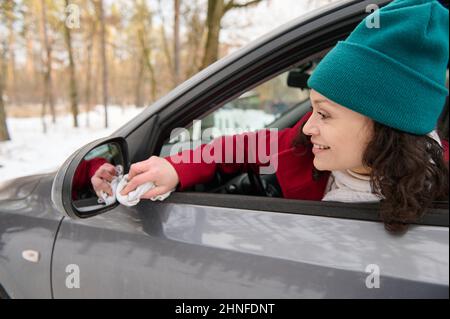 Eine junge Fahrerin sitzt auf dem Fahrersitz ihres Autos und wischt den Seitenspiegel des Fahrers, während sie während der Fahrt bei einer verschneiten Waldlandung anhält Stockfoto