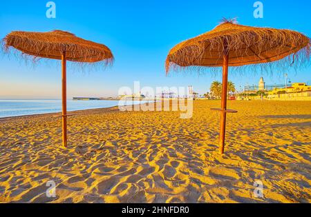 Die wunderschöne Sandküste des Mittelmeers mit Strohsonnenschirmen und dem Hafen von Malaga im Hintergrund, Malagueta-Strand, Malaga, Spanien Stockfoto