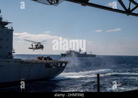 PHILIPPINISCHES MEER (Feb 15, 2022) ein MH-60s Sea Hawk Hubschrauber vom Helicopter Sea Combat Squadron (HSC) 25 führt eine vertikale Auffüllung zwischen dem Flottennachfüller USNS Yukon (T-AO 202) und dem amphibischen Transportschiff USS Green Bay (LPD 20) durch, rechts, Während einer Auffüllung-auf-See zwischen den beiden Schiffen und dem nach vorne eingesetzten amphibischen Angriffsschiff USS America. America, das führende Schiff der America Amphibious Ready Group, arbeitet zusammen mit der 31st Marine Expeditionary Unit im Verantwortungsbereich der US-Flotte für 7th, um die Interoperabilität mit Verbündeten und Partnern zu verbessern Stockfoto