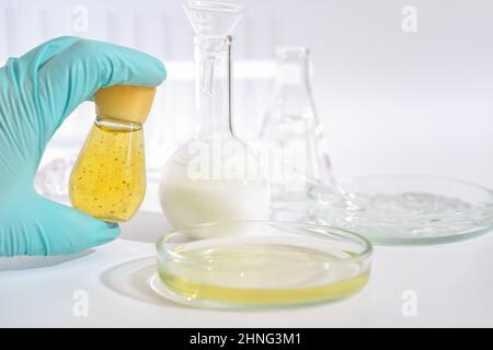 Laborant Hand in medizinischen Handschuh hält eine Flasche kosmetisches Produkt neben einer Petrischale und Glaswaren. Forschung und Prüfung von Kosmetik. Sicherheit und Qualitätskontrolle der Inhaltsstoffe. Stockfoto