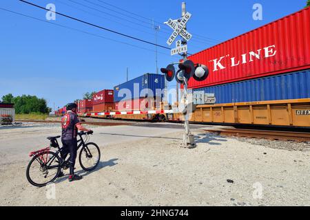 Bartlett, Illinois, USA. Ein einsamer Radfahrer wartet an einer Bahnüberfahrt auf einen intermodalen Güterzug der Canadian National Railway. Stockfoto