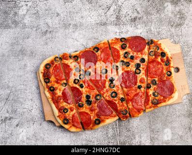 Rechteckige Pizza in Scheiben mit Pepperoni, Käse und Oliven auf einem rechteckigen Holzbrett auf dunkelgrauem Hintergrund. Draufsicht, flach liegend Stockfoto