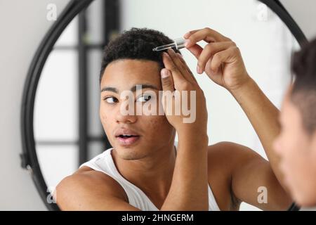 Junger afroamerikanischer Kerl, der Serum für das Haarwachstum im Badezimmer verwendet Stockfoto