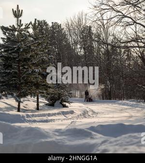 Winterszene mit Schnee auf der Straße und Bäumen und einem Mann, der einen Schneefräsen benutzt, um nach einem Sturm neu gefallenen Schnee von der Auffahrt zu entfernen. Stockfoto