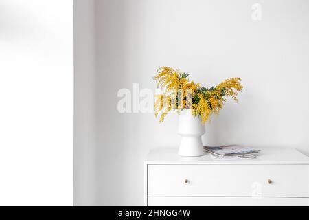 Vase mit Mimosenblüten und Zeitschriften auf Kommode in der Nähe einer hellen Wand Stockfoto