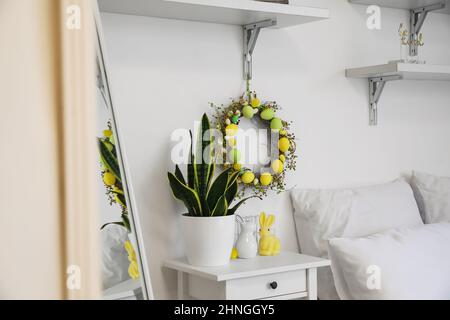 Tisch mit Zimmerpflanze, Osterkaninchen und Kranz, der an einer hellen Wand im Schlafzimmer hängt Stockfoto