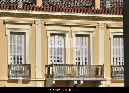 Alte neoklassizistische Hausfassade mit hellgrauen Fensterläden aus Holz, einem Lehmziegeldach und Balkonen mit Eisengeländer in Nafplio, Griechenland. Stockfoto