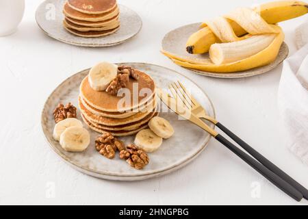 Ein Stapel dicker Pfannkuchen auf einem Keramikplatte mit Bananen und Nüssen auf einem weißen Tisch. Das Konzept eines leckeren Frühstücks oder Snacks Stockfoto