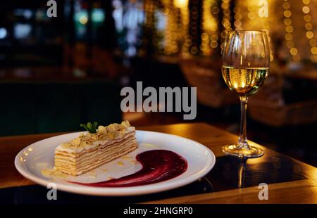 Ein Glas Weißwein und eine italienische Napoleon-Torte und Erdbeersirup in die weiße Keramikplatte auf einem Restauranttisch mit Beleuchtung reflecte Stockfoto