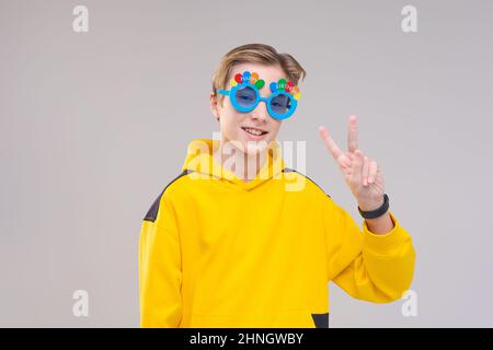 Kaukasischer Kerl in gelbem Sweatshirt und Brille mit Aufschrift Geburtstag posiert glücklich auf grauem Hintergrund, zeigt eine Geste mit der Hand. Freude und Spaß an Ihrem Geburtstag Stockfoto