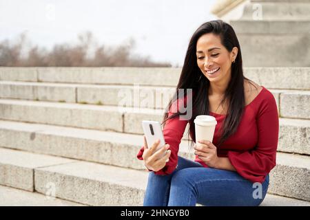 Fröhliche hispanische Frau sitzt auf der Treppe mit einer Tasse Kaffee und mit dem Smartphone zum Chatten Stockfoto