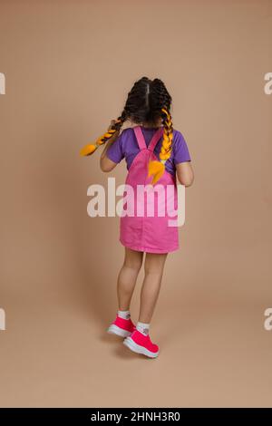 Hinten ein energisches, kleines Mädchen mit zwei Kanekalon-Zöpfen in gelber Farbe, springen in rosa Jumpsuit, lila T-Shirt und rosa Sneakers auf beige Stockfoto