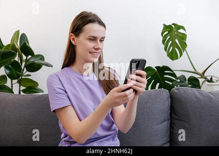 Glücklich lächelnd entspannte junge Frau hält Smartphone mit mobilen Apps lustiges Video ansehen Lachen sitzen auf bequeme Couch, faule Dame Spaß beim Chatten Stockfoto