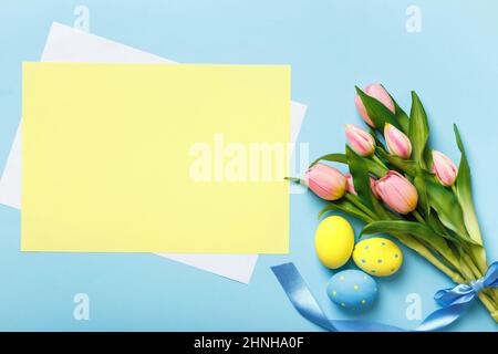 Osterkomposition. Eier Blumen Tulpen auf einem blauen Hintergrund. Leeres Blatt Papier mit Platz für Text und Kopierer Stockfoto