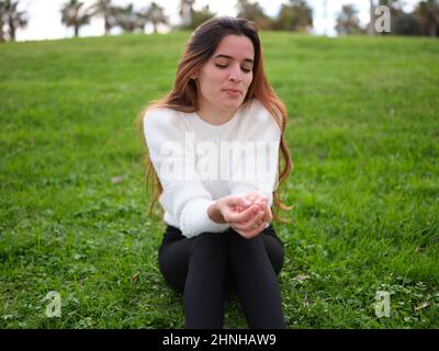 Eine schüchterne junge Frau, die mit geschlossenen Augen auf dem Gras im Park sitzt und ihre Hände an ihren Beinen zusammenhält. Stockfoto