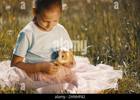 Das Mädchen sitzt auf dem Gras und füttert ihr Meerschweinchen mit einer Dandelionenblume. Stockfoto