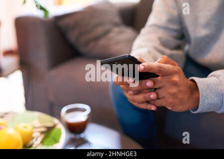 Die Hände eines Mannes tippen auf einem Smartphone, während er auf der Couch sitzt Stockfoto