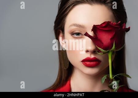 Nahaufnahme einer jungen Frau in der Nähe der roten Rose, isoliert auf grau Stockfoto
