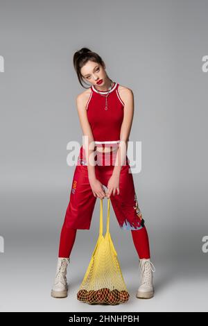 In voller Länge trendige junge Frau in rot sportlichen Outfit mit String-Tasche mit Äpfeln auf grau Stockfoto