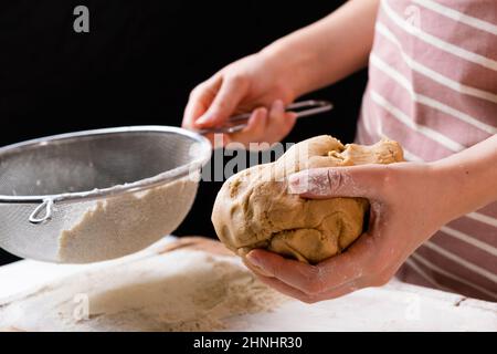 Nahaufnahme Frau siebt Weizenmehl durch ein Sieb in eine Rührschüssel zur Teigvorbereitung für Kuchen und Backwaren auf schwarzem Hintergrund. Den Teig für hausgemachtes Gebäck kneten. Stockfoto