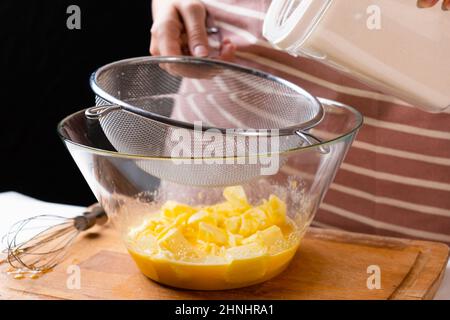 Nahaufnahme Frau siebt Weizenmehl durch ein Sieb in eine Rührschüssel zur Teigvorbereitung für Kuchen und Backwaren auf schwarzem Hintergrund. Den Teig für hausgemachtes Gebäck kneten. Stockfoto