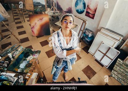 Blick auf einen Künstler, der die Kamera anschaut. Fantasievolle junge Malerin, die mit gekreuzten Armen vor einer Leinwand steht. Kreativer junger woma Stockfoto