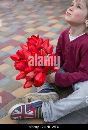 Netter Junge 4-5 Jahre alt hält einen großen Strauß roter Tulpen und schaut mit Hoffnung auf. Muttertag. Frühlingsblumen für Mama, Freundin. Symbol der Liebe, Stockfoto