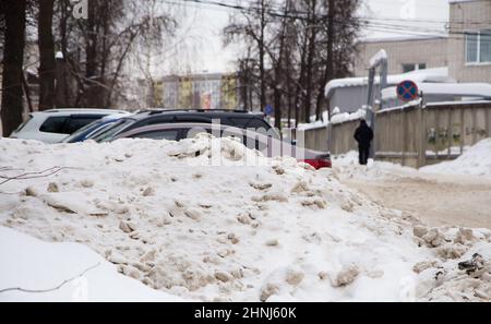 Schmutzige Schneeverwehung an der Straße vor dem Hintergrund einer Stadtstraße mit Autos. Auf der Straße liegt schmutziger Schnee in hohen Haufen. Urbane Winterlandschaft. Wolkiger Wintertag, weiches Licht. Stockfoto