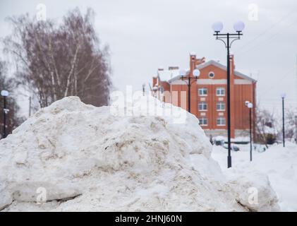 Eine massive Schneeverwehung an der Straße vor dem Hintergrund von Stadthäusern und Bäumen. Auf der Straße liegt schmutziger Schnee in hohen Haufen. Urbane Winterlandschaft. Wolkiger Wintertag, weiches Licht. Stockfoto
