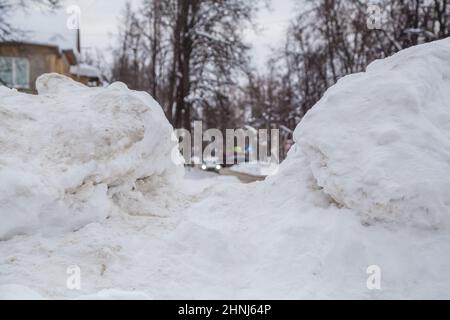 Eine breite, große Schneeverwehung an der Straße vor dem Hintergrund einer Stadtstraße. Auf der Straße liegt schmutziger Schnee in hohen Haufen. Urbane Winterlandschaft. Wolkiger Wintertag, weiches Licht. Stockfoto