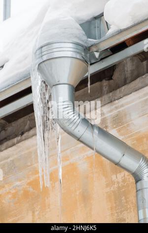 Lange Eiszapfen hängen an einem Abflussrohr am Rand des Daches. Vor dem Hintergrund der Mauer eines alten Backsteinhauses. Große Kaskaden, sogar schöne Reihen. Wolkiger Wintertag, weiches Licht. Stockfoto