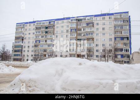 Eine riesige, große Schneeverwehung an der Straße vor dem Hintergrund der Stadthäuser. Auf der Straße liegt schmutziger Schnee in hohen Haufen. Urbane Winterlandschaft. Wolkiger Wintertag, weiches Licht. Stockfoto