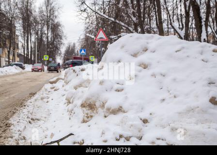 Eine große Schneeverwehung am Straßenrand vor dem Hintergrund einer Stadtstraße. Auf der Straße liegt schmutziger Schnee in hohen Haufen. Urbane Winterlandschaft. Wolkiger Wintertag, weiches Licht. Stockfoto