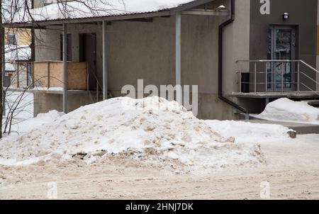 März große Schneeverwehung durch die Straße vor dem Hintergrund der Stadthäuser. Auf der Straße liegt schmutziger Schnee in hohen Haufen. Urbane Winterlandschaft. Wolkiger Wintertag, weiches Licht. Stockfoto