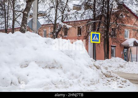 Eine mächtige Schneeverwehung an der Straße vor dem Hintergrund von Stadthäusern und Bäumen. Auf der Straße liegt schmutziger Schnee in hohen Haufen. Urbane Winterlandschaft. Wolkiger Wintertag, weiches Licht. Stockfoto