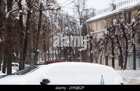 Eine lockere Schneeverwehung von Autos an der Straße vor dem Hintergrund einer Stadtstraße. Auf der Straße liegt schmutziger Schnee in hohen Haufen. Urbane Winterlandschaft. Wolkiger Wintertag, weiches Licht. Stockfoto