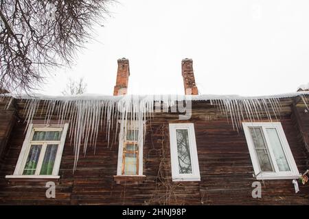 An der Dachkante hängen transparente Eiszapfen. Vor dem Hintergrund der Holzwand des alten Hauses. Große Kaskaden, sogar schöne Reihen. Wolkiger Wintertag, weiches Licht. Stockfoto