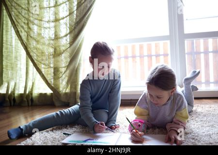 Zwei kleine niedliche Mädchen zeichnen ein Album, das auf dem Boden neben einem großen Fenster liegt Stockfoto