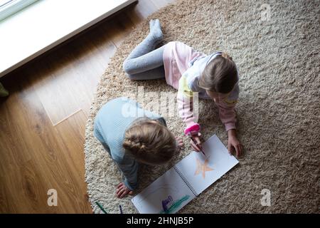 Zwei kleine niedliche Mädchen zeichnen ein Album, das auf dem Boden neben einem großen Fenster liegt Stockfoto