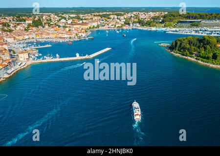 Yacht segelt in Richtung Meeresbucht mit Rovinj Stadt auf Halbinsel. Kroatische Stadtgebäude mit roten Dächern, umgeben von Bäumen und Adria. Luftpanorama Stockfoto