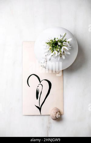 Strauß frischer Schneeglöckchen Frühlingsblumen in weißer Porzellanvase mit Schneckenschale und handgezeichneter Schneeglöckchen auf weißem Marmortisch. Frühlingszeit komposit Stockfoto