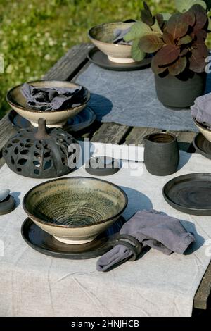 Rustikale Tischgestaltung draußen im Garten mit leerem handwerklichem Keramikgeschirr, schwarzen Tellern und rauen Schüsseln, Kürbisdekorationen, auf Leinentischdecke Stockfoto