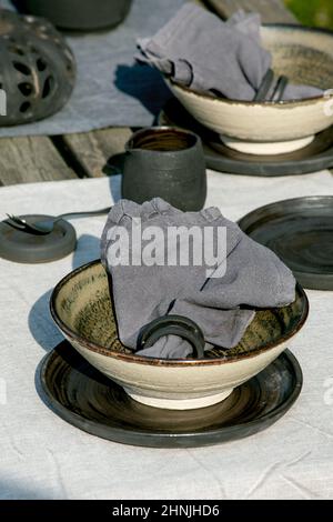 Rustikale Tischgestaltung draußen im Garten mit leerem handwerklichem Keramikgeschirr, schwarzen Tellern und rauen Schüsseln, Kürbisdekorationen, auf Leinentischdecke Stockfoto