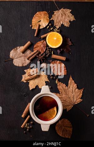 Heißer Glühwein im Keramiktopf mit Gewürzen, Orange und Herbstblättern auf schwarzem Holzboden. Flach liegend. Gemütliche warme Getränke Stockfoto